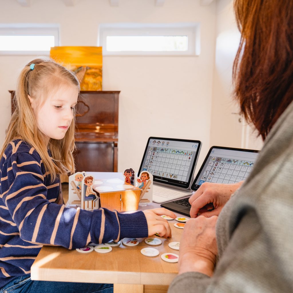 Ein Mädchen während einer logopädischen Bahndlung. Sie spielt ein Brettspiel und neben ihr sind zwei Tablets. Bei ihr ist die Logopädin.